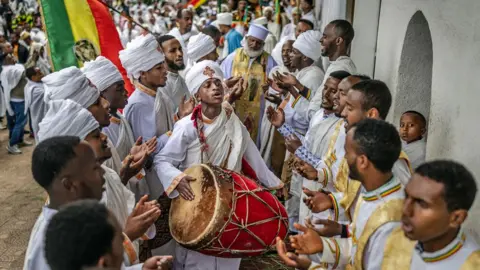 लुईस टाटो/एएफपी/गेटी इमेजेस ऑर्थोडॉक्स डिकन्स आणि एक याजक पांढ white ्या धार्मिक कपड्यांचा एक तरुण माणूस आहे जो अ‍ॅडिस अबाबा, इथिओपियर अँटो सेंट रागुएल नवीन वर्षाच्या उत्सव दरम्यान एक मोठा लाल ड्रम गातो - गुरुवारी 11 सप्टेंबर 2025
