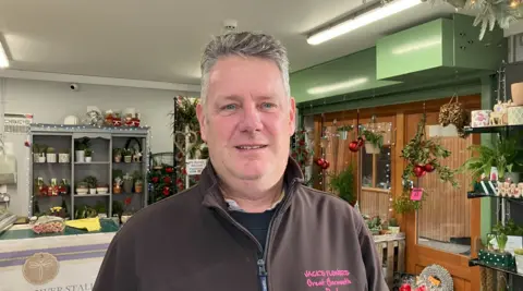 A grey-haired man dressed in a dark brown fleece looks at the camera with a flower stall behind him.
