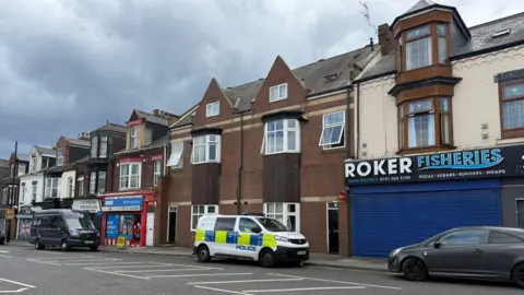 Roker Avenue in Sunderland. There are a row of terraced houses, takeaways and an off-license. There is a police van parked outside the houses.