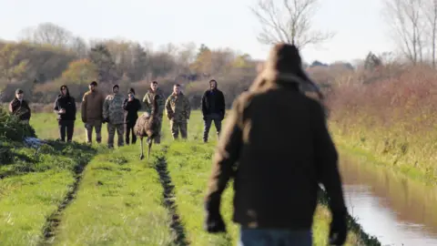 Cleethorpes Wildlife Rescue A person in a brown coat and jeans wearing a brown hat walking towards a group of people in camoflauged clothing who are crowding around an emu on the loose. They are in a field with green grass.