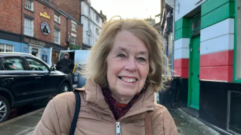 Eleri facing the camera and smiling, she's wearing a brown winter jacket and has a red scarf on as well. Behind her you can see Castle Gates, with the Bulls Head pub visible over one shoulder. It's a cold wintery day, with low sunlight just coming through the clouds. 