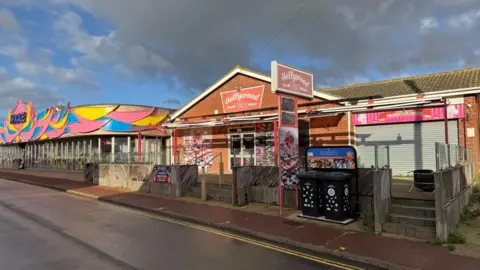 Andrew Turner/BBC Hollywood Diner is a 1980s red brick building with a gabled end facing Beach Road, large windows, a canopy and signs outside, where there is a terrace outdoor dining area. The property is empty. Next to it is the brightly coloured facade of the Mirage amusement arcade, which has a wraparound glass door.