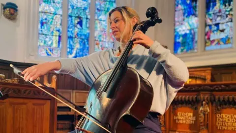 Woman with blonde hair and wearing grey top, playing a dark wood cello, in a chapel setting with stained glass windows in the background