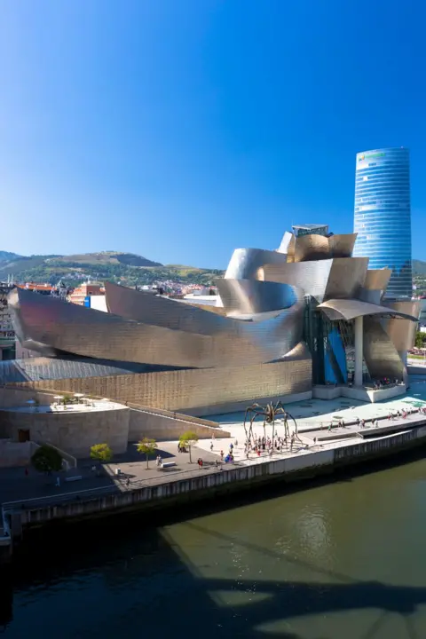 Tim Graham/Getty Images A view of the Guggenheim Museum in Bilbao, Spain. It shows the building's unique silver material, glistening in bright sunshine, around its iconic curvature.
