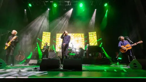 Getty Images Deacon Blue playing on stage with green and white lights shining down on them. The stage is illuminated, mainly in green. 