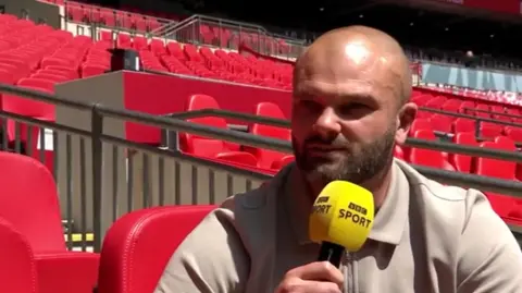 A man wearing a cream shirt sat in the stands at Wembley. He is surrounded by red seat and is holding a yellow BBC Sport microphone