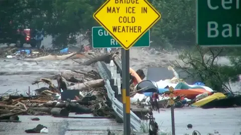 A raging river rushes over a bridge that is strewn with debris.