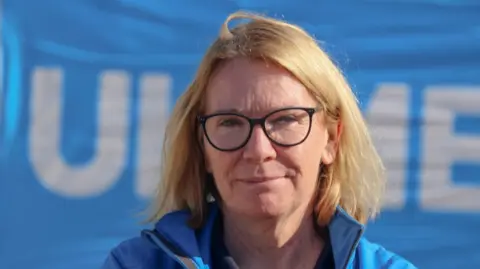 A woman with blonde hair and glasses, wearing a blue jacket