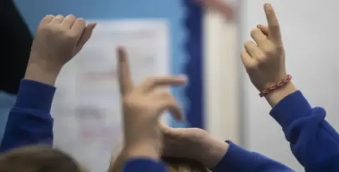 Kids wearing blue school jumpers with their hands in the air.
