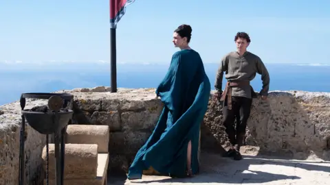 Universal Pictures Anne Hathaway as Penelope (Odysseus's wife) in centre of image in blue gown looking to the side and Tom Holland as their son Telemachus leaning on wall in khaki top and dark trousers.
Blue skies behind
