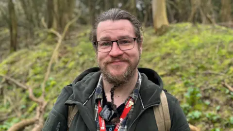 BBC A man standing in a forest in front of a mossy bank with a beard and wearing glasses. He wears a green jumper and is carrying a rucksack. 
