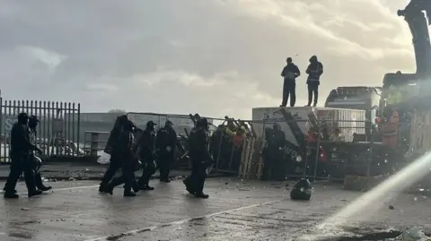 RTE A number of Garda officers dressed in black riot polices-style uniforms move towards a barricade of pallets and wire fencing which has two men standing on top of a container behind it. A lorry with a loading arm can also be seen further back.