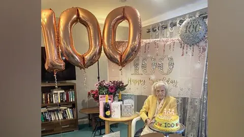 Elderly woman in a yellow blouse and jacket wears cream slacks and cream flat shoes. She is seated and in front of her is a large yellow and cream-iced birthday cake with 100 in piped icing on it. On another table are cards, champagne and balloons and a banner with 100 on them.