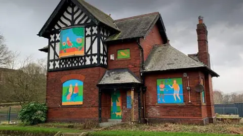 Bitts Park Lodge is a red brick detached building with a Tudor black and white cladding on its front. Its windows are boarded up with decorative paintings on them. 