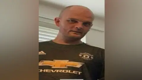 Hertfordshire Constabulary Gavin Hubbard, who has a shaven head and stubble, wearing an old Manchester United away shirt. He is looking down at the camera. 
He is standing in front of a window which has a venetian blind that is closed.