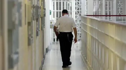 PA Media a prison officer in uniform walking away from the camera along a prison wing of cells