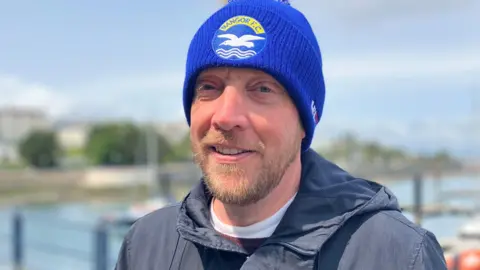 A man is standing in a path in front of the sea. He's wearig a blue football hat and a grey coat.