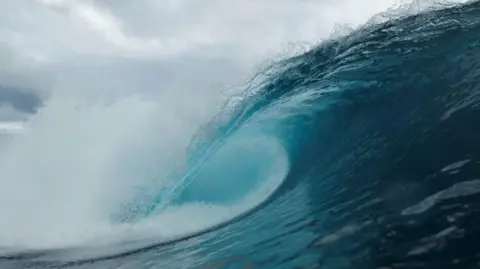 The crescent of a wave is seen crashing over. The wave is dark blue and lighter towards the tip of the wave and there is white spray coming away from the wave. The sky is cloudy.