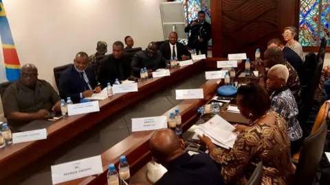 Monusco/X Rebels and UN representatives at the meeting in Goma. They sit opposite each other at long, parallel desks made of dark wood and each has a name card in front of their seat.