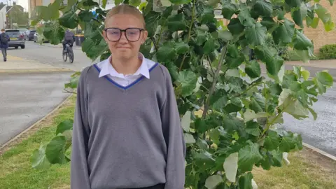 Bedford Giving Ellie, standing outside, by a bush, smiling and looking straight at the camera, with fair hair tied back, wearing glasses, a grey jumper and skirt underneath it, people are behind her in the distance.