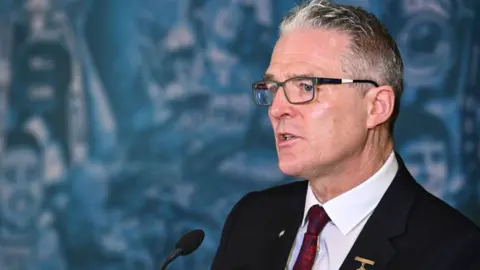 Jarlath Burns, he is wearing a black suit with a red tie and glasses. He has dark grey hair.