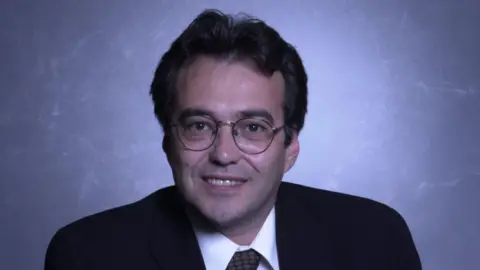 Phil Woolas, wearing a dark suit, tie and glasses, smiles straight in a posed photo from his time as an MP.
