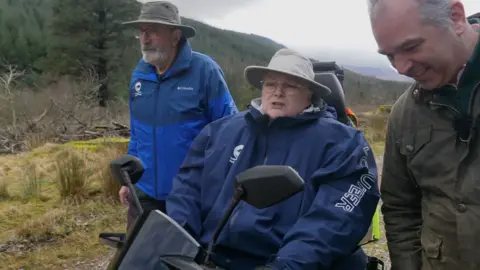 A woman using a mobility scooter with a dark blue jacket in the middle, travelling along a wide gravel track. Two men walk beside her, one on each side. The track runs through open countryside with grassy ground, scattered trees, and forested slopes rising on either side. Cloudy sky overhead.