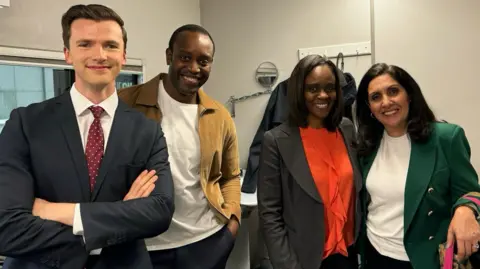 Dan Heard in a suit and tie. Beside him, standing in a row, iare Gamal Fahnbulleh, Gillian Joseph, and Maryam Moshiri. They are all wearing smart jackets and smiling as they look at the camera.