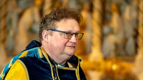 James Garrard is looking sideways and has short, curly brown hair. He is wearing blue-rimmed spectacles and blue and gold robes