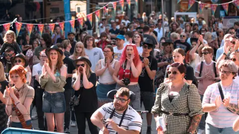 City of Lincoln Council Scores of people, with their faces towards the camera lens, watch an outdoor stage performance at Lincoln Live. Some people are wearing summer clothes and sunglasses as they clap their hands and others shout. Two rows of red, blue and yellow bunting can be seen overhead.