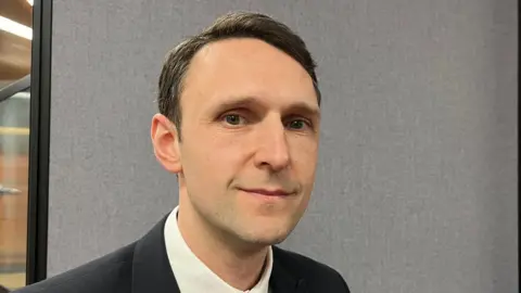 BBC Professor David Hughes is sitting in front of a grey wall and looking directly at the camera. He is wearing a navy suit, tie and white shirt. He has short black hair.