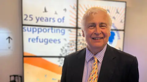 Jon Wright/BBC Sir Nick Young wearing a suit and tie stands in front of a banner saying 25 years of supporting refugees