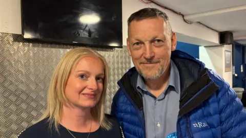 Martin Heath/BBC Craig Stanbury with short brown hair and beard, wearing a blue and black anorak and grey shirt, standing next to Claire Hunt, with long blonde hair and wearing a black T-shirt. They are both looking at the camera in a bar-like room with a TV on the wall behind them.