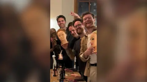 Four men smiling at the camera and gesturing to plastic heads with plaited hair.