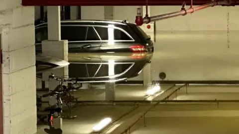 A flooded car park with the water level up to the break lights of two parked cars in the background. The rear brake lights are reflected in the water