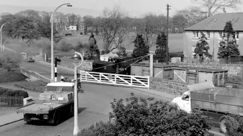 Don Martin A level crossing, which crossed the road between Kirkintilloch and Lenzie near Woodhead Park. A train is passing through while a flat bed truck waits to cross