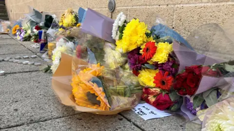Jon Wright/BBC Bunches of flowers, candles and tealights are placed against a wall. Cans of drink are left too.