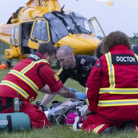 Lincs and Notts Air Ambulance A doctor and a paramedic, dressed in red jackets with hi-vis stripes, and a pilot wearing a black shirt, kneel on a green field as they treat a patient with medical equipment. In the background is a yellow helicopter, its doors open. 