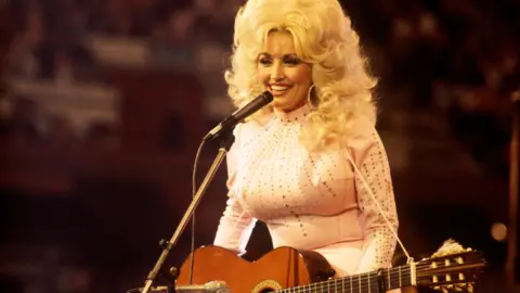 David Redfern/Redferns/Getty Images Dolly Parton in 1976 smiling on stage with large voluminous blonde hair, big hoop earrings and a white long top with gems on it. She is holding an acoustic guitar and a microphone is in front of her.