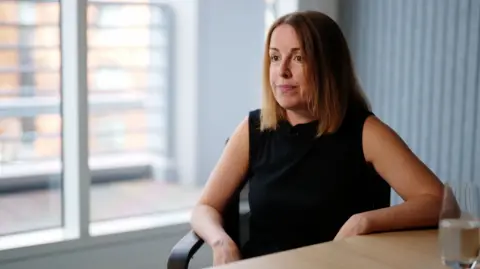 Adam Walker/BBC Tessa Gregory, a woman with brown, shoulder-length hair, wearing a black top and sitting at a desk with a glass of water in front of her and a window with a view of the street behind her.