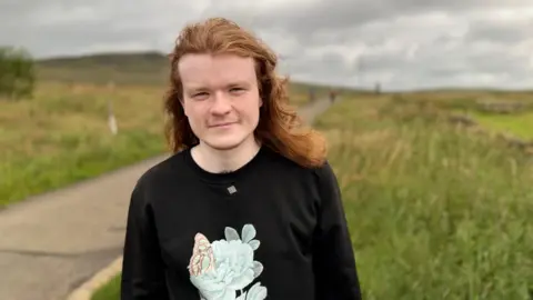 A man with long ginger hair, wearing a black top. He is standing in a field with a grey sky above.