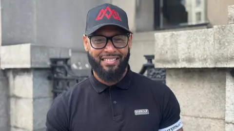 Miguel Doforo has a black beard and is wearing dark framed glasses. He is wearing a black baseball cap and a navy blue t-shirt and is stood outside St Helens library and smiling at the camera.
