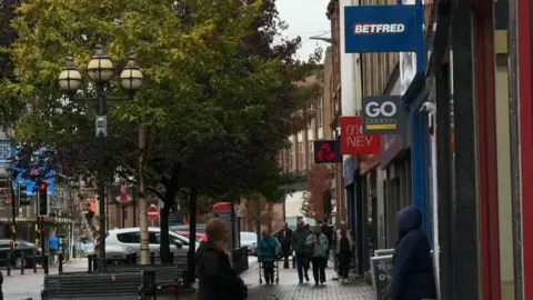People walk down an English street in Carlisle. On the right is a row of shops with signs visible from the buildings. In order of precedence, these are Betfred, Go Outdoors, Virgin Money and NatWest. There are trees in full bloom and an ornate lamppost topped with three globe-shaped lights. There are benches and cars parked at the end of what looks like a pedestrian street. People are dressed in coats, indicating a cold day.