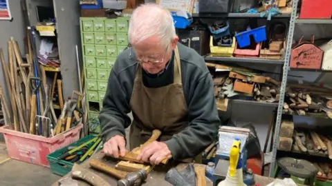 A man works in a workshop. He has grey hair and glasses and is looking down at woodwork he is working on. There are tools in the background.