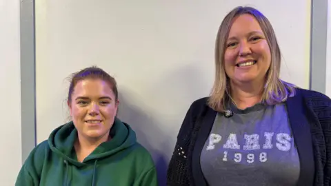 Two women, one wearing a green hoodie with tied-back brown hair and one with shoulder-length blonde hair wearing a T shirt and cardigan.