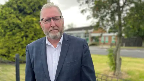Doug Beattie has grey hair and a grey beard. He is wearing wired framed, rectangular frames, a navy jacket, white shirt and navy tie. 
