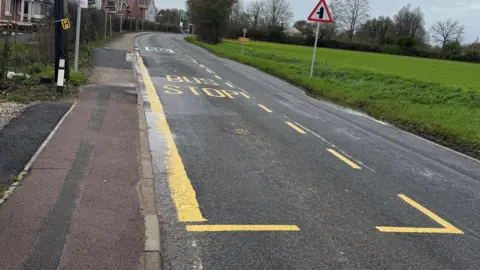 Nadira Tudor/BBC A bus stop outlined by yellow markings painted on a road, which is flanked by a field on one side and houses on the other.
