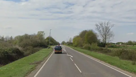 A van is driving on the A142 away from the camera. There are trees, bushes and fields either side of the single carriageway road. There is blue skies with white cloud.