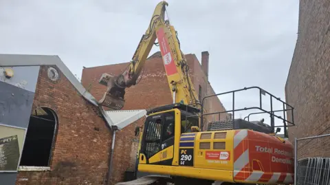 A bright yellow demolition vehicle tears the corrugated metal roof from the top of the building.
