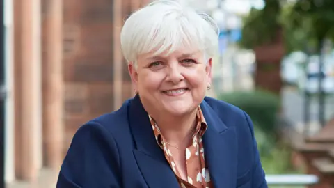 Head and shoulders photo of Angela O'Hagan. She is outside but the background image is blurred out. She is looking directly at the camera, wearing a blue blazer jacket and a brown blouse with white dots. She has grey hair which is long on top but does not go below the ears at the front. 